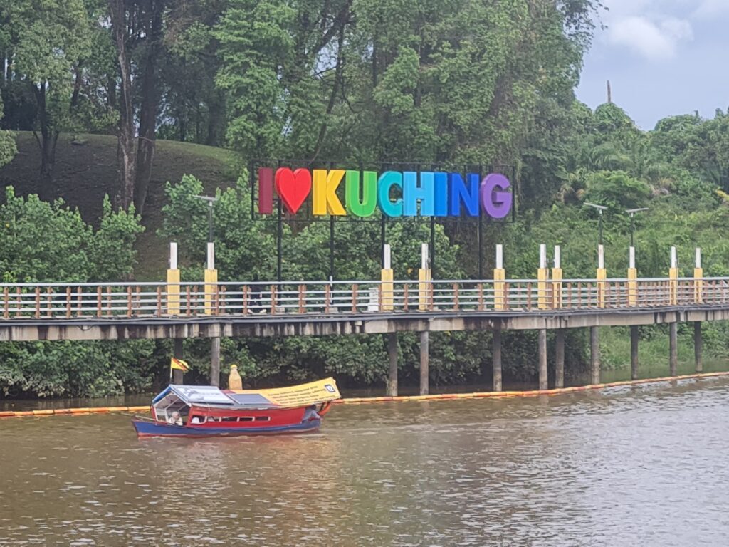 The colorful 'I Love Kuching' sign on the Sarawak Riverfront with a traditional tambang boat passing by.