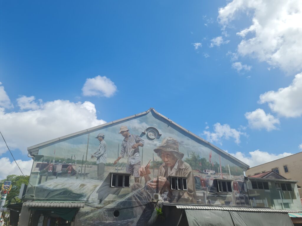 Large street art mural in Kuching depicting a fisherman mending nets on a blue sky day.