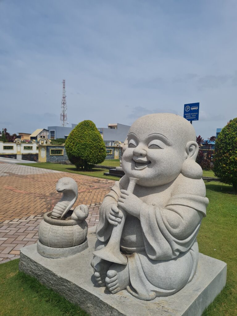 Stone statue of a smiling Buddha playing a flute next to a cobra statue in the garden.