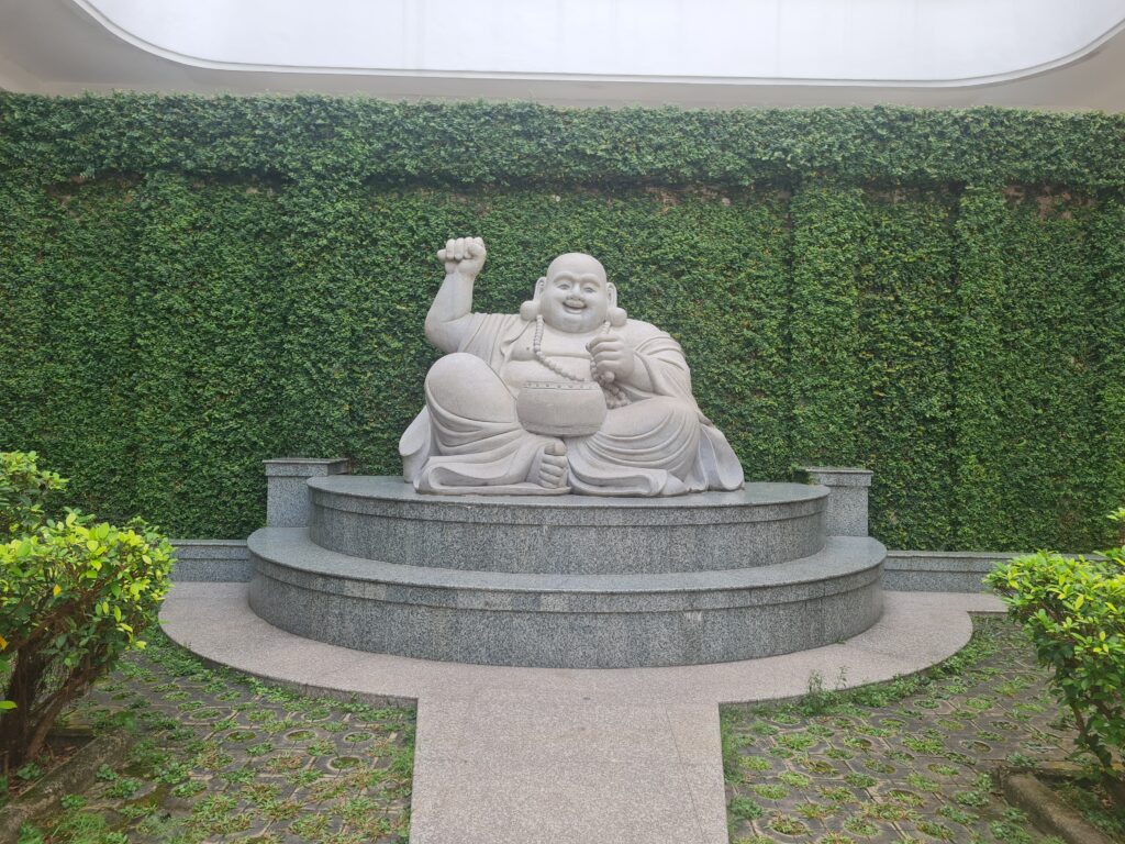 Stone statue of the Laughing Buddha Maitreya sitting in a lush green garden at the monastery.